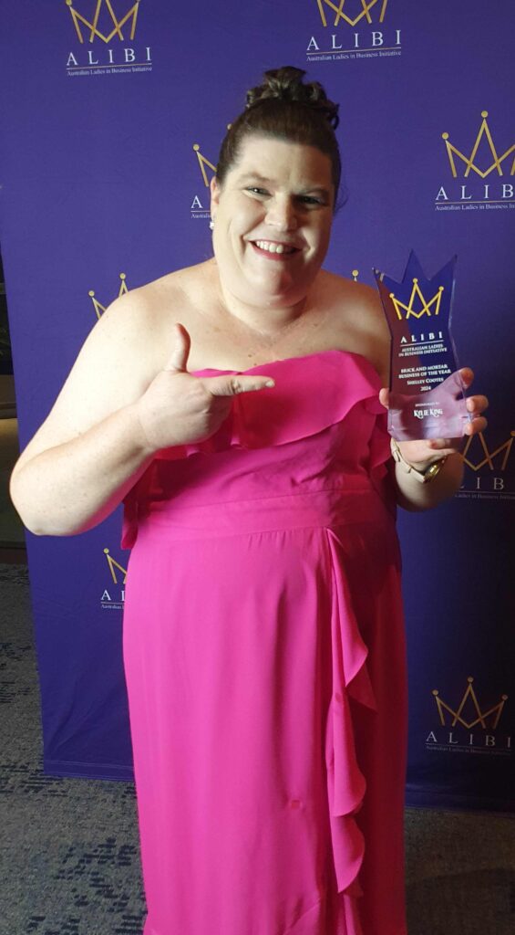 Shelley in a formal pink dress pointing to the Brick and Mortar Business of the Year Award which is a beautiful purple trophy